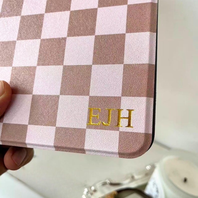 May include: A close-up of a case with a pink and brown checkered pattern. The initials "EJH" are displayed in gold lettering on the lower right corner of the case. The case is held by a hand.