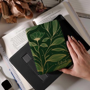 May include: A dark green book cover features a gold floral design and the name "Astride". The cover is held over a black book stand, with open books visible in the background.