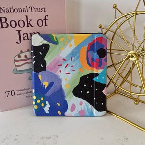 以下が含まれることがあります： 青、黄、黒、赤などの鮮やかな色使いの抽象的なアートが施された、正方形のケース。電子書籍リーダーやタブレット用かもしれません。モダンで芸術的な雰囲気のデザインです。ケースの隣には本と金色の金属製の装飾品があります。