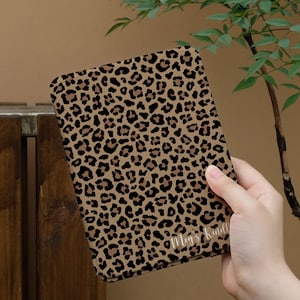 Peut inclure: Une housse de Kindle avec un imprimé léopard beige et noir. La housse présente un motif répété de taches noires sur un fond beige. Les mots "Mey's Kindle" sont imprimés en blanc en bas à droite.