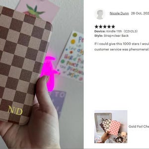 May include: A brown and tan checkerboard Kindle case with gold initials "NID". The case has a strap and clear back. A gold charm hangs from the bottom. The case is held in a hand.