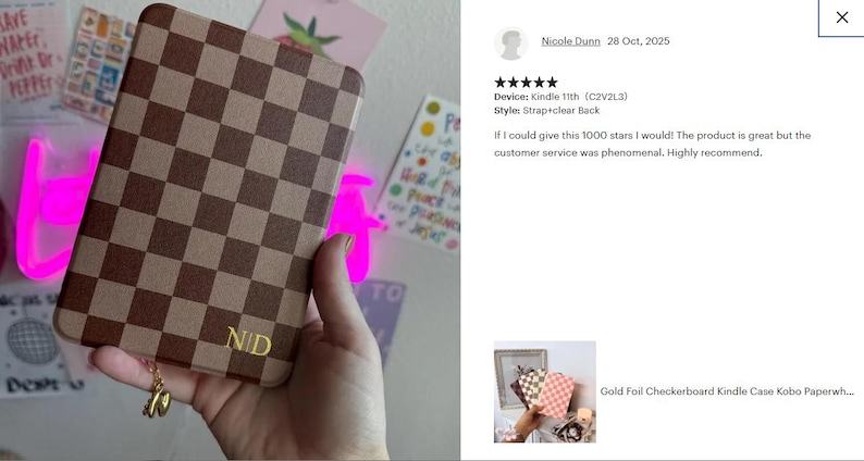 May include: A brown and tan checkerboard Kindle case with gold initials "NID". The case has a strap and clear back. A gold charm hangs from the bottom. The case is held in a hand.