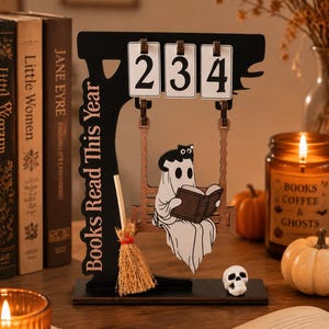May include: A black wooden countdown sign with the text "Books Read This Year" featuring a white ghost sitting on a swing reading a book with a small black cat on its head. Three white flip cards display the number 234, and a miniature broom is attached.