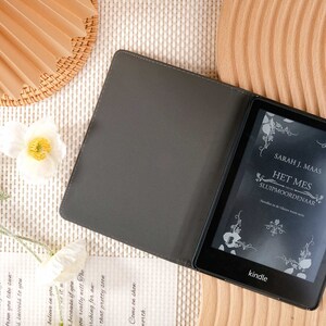 May include: A black Kindle e-reader with a gray cover. The screen displays the title of a book in Dutch, "Het Mes van de Sluipmoordenaar" by Sarah J. Maas.