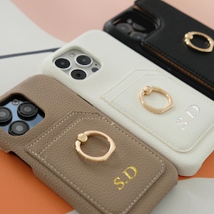 May include: Close-up of several phone cases in various colors: taupe, white, and black. Each case features a cardholder and a gold ring holder. The cases are personalized with the initials "S.D."