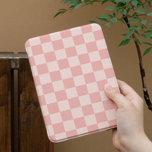 May include: A pink and white checkered pattern tablet case. The case is rectangular with rounded corners. The case is held in front of a brown background with green leaves.