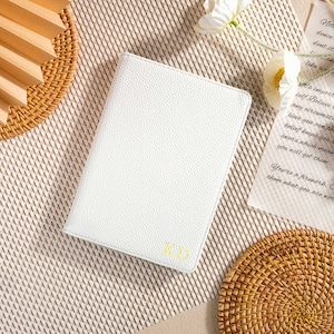May include: A white leather-bound journal with the initials "KD" embossed in gold on the front cover. The journal is lying on a woven straw mat.
