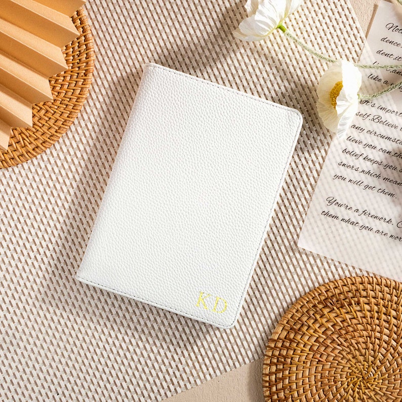 May include: A white leather-bound journal with the initials "KD" embossed in gold on the front cover. The journal is lying on a woven straw mat.