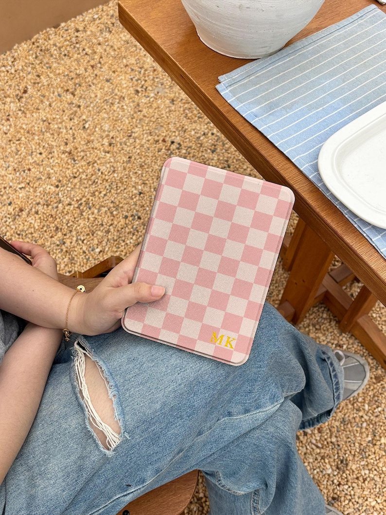 May include: A pink and white checkered tablet case with the initials "MK" in gold. The case is held by a person wearing blue jeans. The background includes a wooden table and a gravel surface.