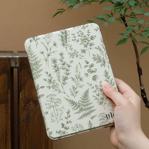 May include: A rectangular e-reader case featuring an off-white background with a detailed botanical pattern of green ferns, herbs, and various leaves. The case has rounded corners and a partial text element visible in the lower right corner.