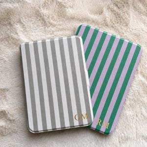 May include: Two rectangular e-reader cases with rounded corners. One case features vertical grey and white stripes with the gold monogram GM. The second case features vertical green and lavender stripes with the gold monogram RM.