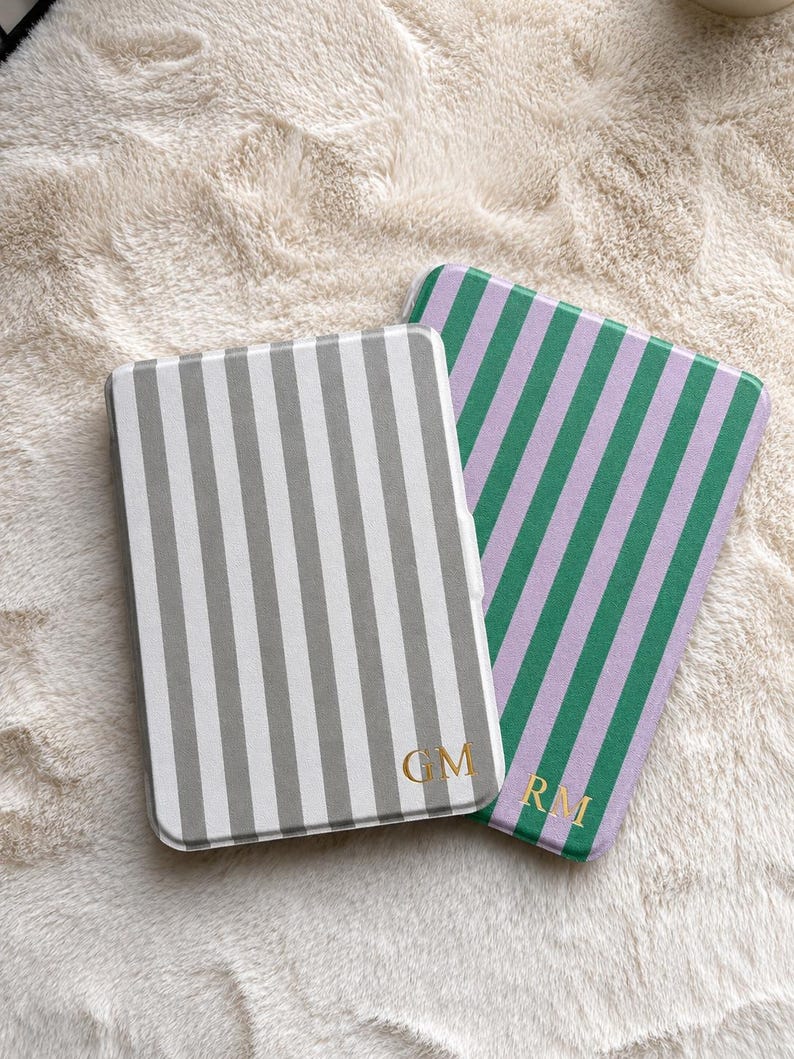 May include: Two rectangular e-reader cases with rounded corners. One case features vertical grey and white stripes with the gold monogram GM. The second case features vertical green and lavender stripes with the gold monogram RM.