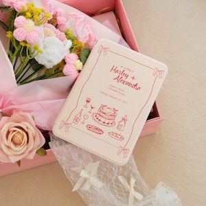 May include: A pink gift box contains a bouquet of crochet flowers, a pink rose, and a light pink rectangular box. The box is decorated with red line drawings of food and drink, and the text "Harley + Alexandra".