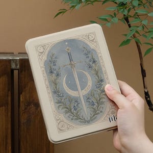 May include: A rectangular e-reader case featuring a cream-colored background with a central illustration of a silver sword pointing downward, framed by blue bell-shaped flowers and a crescent moon against a soft blue, star-dusted oval backdrop.