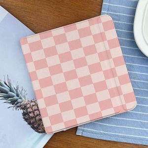 May include: A pink and white checkered tablet case. The case is rectangular with rounded corners and a light pink and white checkerboard pattern. The case is on a wooden surface next to a pineapple and a blue and white striped cloth.