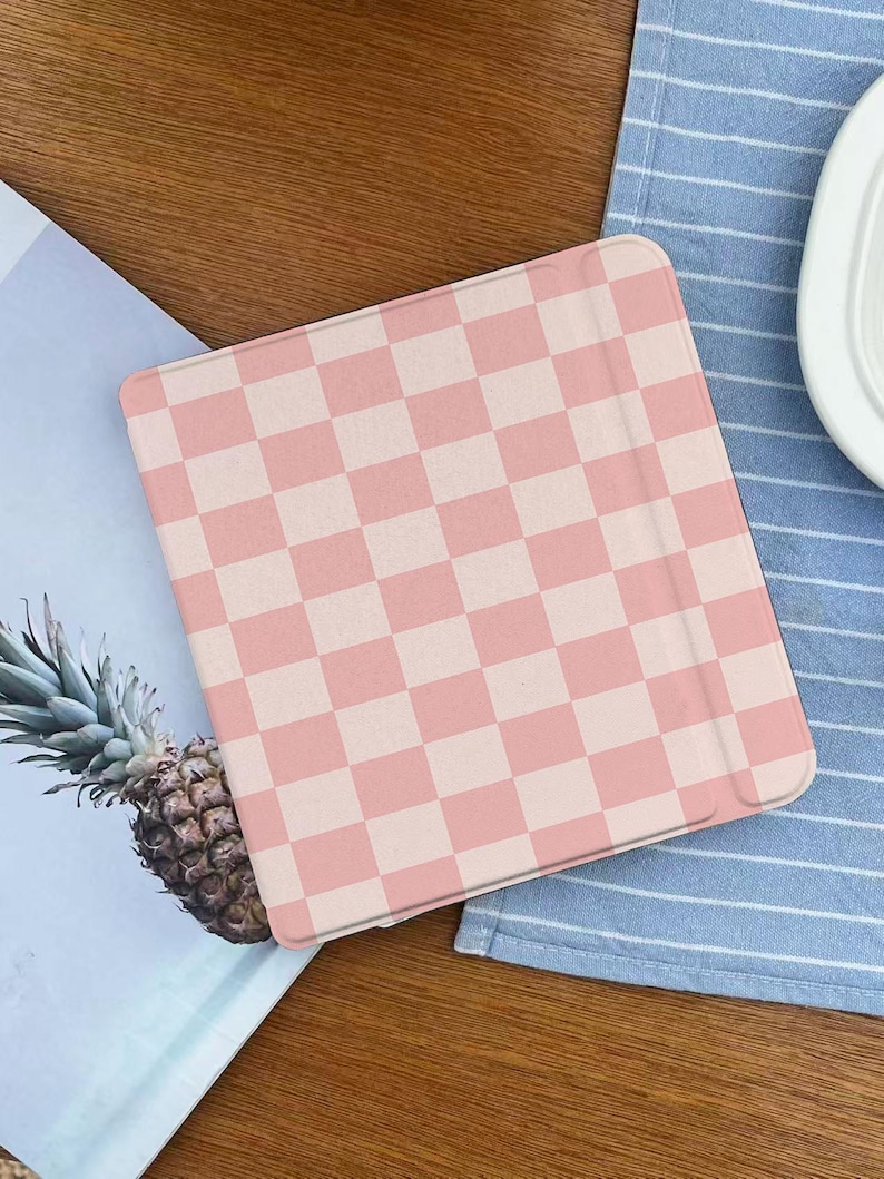 May include: A pink and white checkered tablet case. The case is rectangular with rounded corners and a light pink and white checkerboard pattern. The case is on a wooden surface next to a pineapple and a blue and white striped cloth.