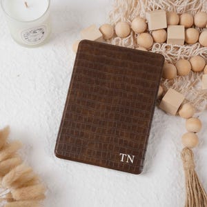May include: A brown, rectangular, leather-textured notebook with the initials "TN" in white. The notebook is surrounded by decorative elements, including a lit candle in a glass jar, wooden beads, and dried flowers.
