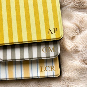 May include: Three rectangular tablet cases with vertical stripes. The top case is yellow and white with the gold embossed letters AP. The middle case is grey and white with the gold letters GM. The bottom case is white, yellow, and blue with LCR.