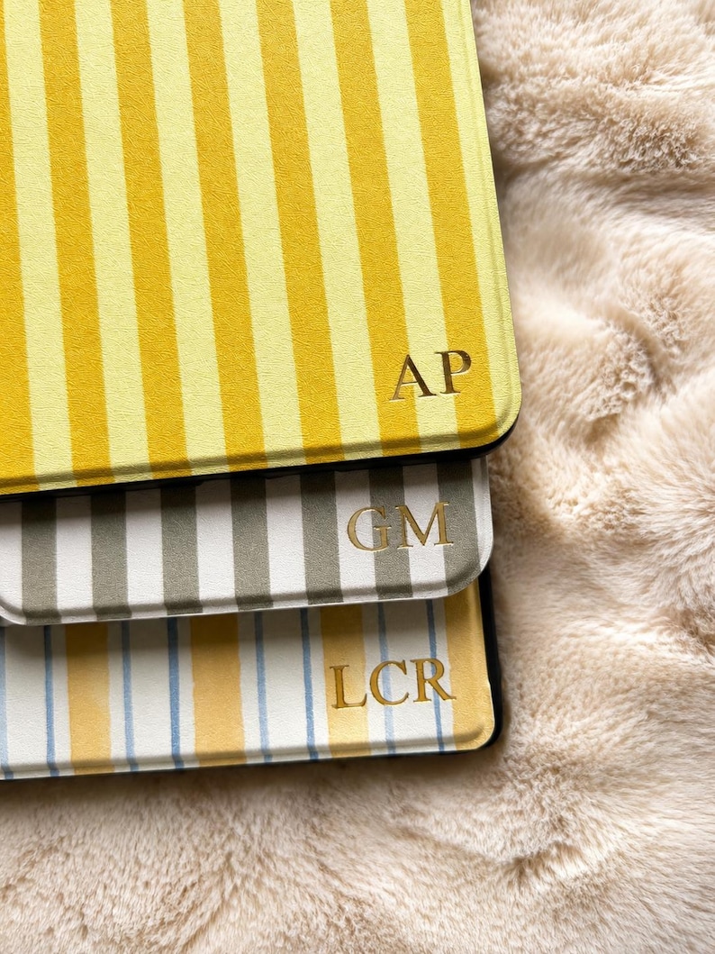 May include: Three rectangular tablet cases with vertical stripes. The top case is yellow and white with the gold embossed letters AP. The middle case is grey and white with the gold letters GM. The bottom case is white, yellow, and blue with LCR.