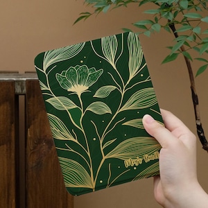 Peut inclure: Une housse de liseuse vert foncé ornée d'une illustration au trait doré représentant une fleur et des feuillages. La housse porte l'inscription "May's Kindle" en police cursive dorée, avec de petits points dorés dispersés sur la surface.