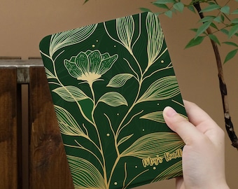 May include: A dark green e-reader cover featuring a gold line art illustration of a flower and foliage. The cover includes the text "May's Kindle" in a gold script font, with small gold dots scattered across the surface of the rectangular device.