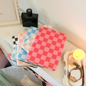 以下が含まれることがあります： ピンク、青、黒、白の市松模様のカラフルな携帯電話ケースのスタック。ケースは白い表面に配置され、背景には香水瓶と火の灯ったキャンドルがあります。