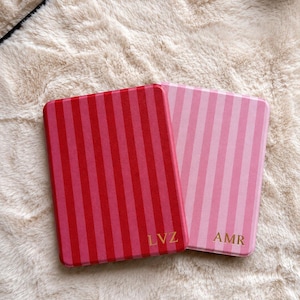 May include: Two rectangular cases with vertical stripes. The left case is dark red and pink with the gold letters LVZ. The right case is light pink and white with the gold letters AMR. Both cases have rounded corners and a smooth, flat surface.
