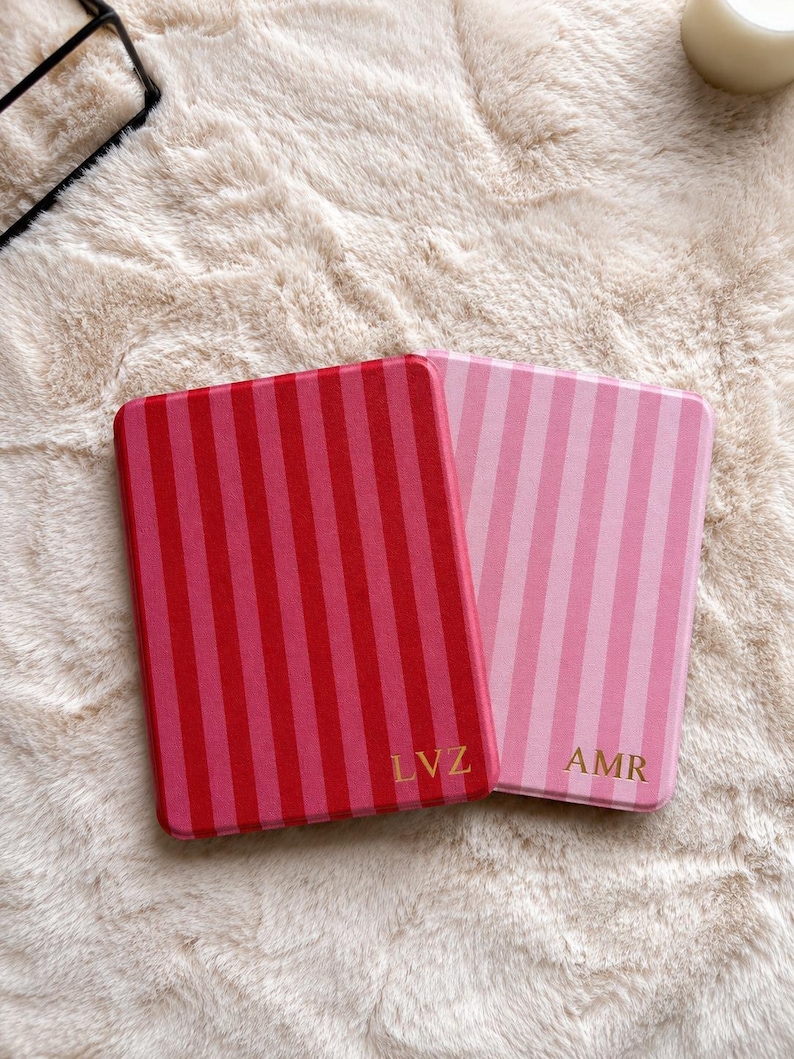 May include: Two rectangular cases with vertical stripes. The left case is dark red and pink with the gold letters LVZ. The right case is light pink and white with the gold letters AMR. Both cases have rounded corners and a smooth, flat surface.