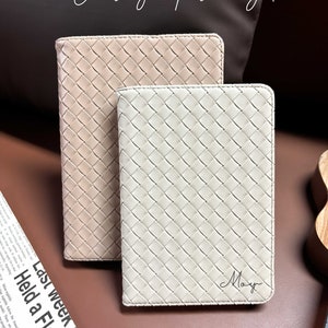 Puede incluir: Dos cuadernos de cuero trenzado, uno beige y otro blanco. El cuaderno blanco tiene el nombre "May" escrito en cursiva.
