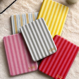 May include: Five rectangular cases with vertical stripes in yellow, grey, pink, red, and blue. Three cases feature gold-colored monogram letters: AMR on the pink case, GM on the grey case, and LVZ on the red case. Each case has rounded corners.