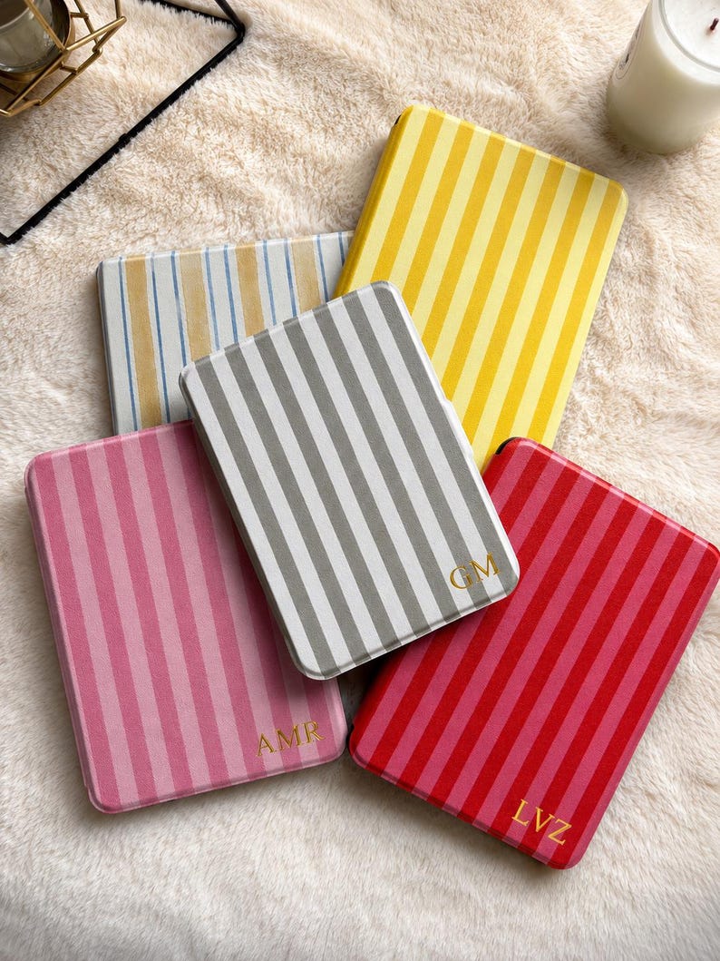 May include: Five rectangular cases with vertical stripes in yellow, grey, pink, red, and blue. Three cases feature gold-colored monogram letters: AMR on the pink case, GM on the grey case, and LVZ on the red case. Each case has rounded corners.