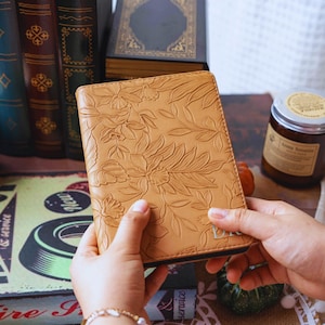May include: A tan leather journal with an embossed floral design. The journal is held open, revealing the interior pages. The cover features a detailed pattern of flowers and leaves. The journal is surrounded by books and a candle.