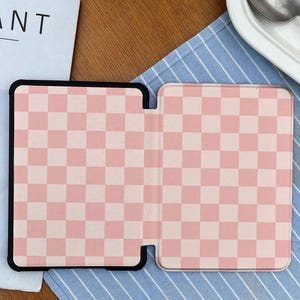 May include: An open tablet case with a pink and white checkered pattern. The case is black around the edges and is lying on a blue and white striped cloth. The word "ANT" is partially visible on a white page.