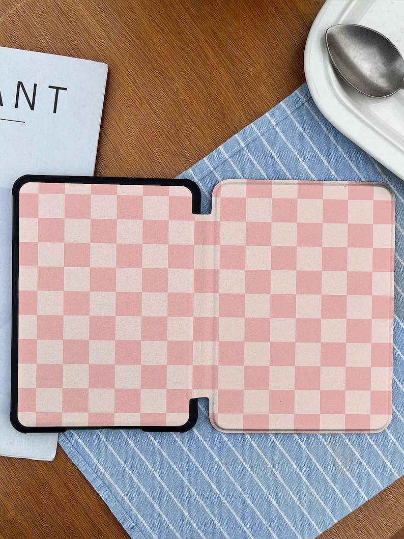May include: An open tablet case with a pink and white checkered pattern. The case is black around the edges and is lying on a blue and white striped cloth. The word "ANT" is partially visible on a white page.
