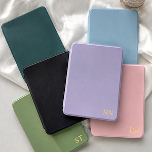 May include: A collection of six rectangular, solid-coloured cases in dark green, light blue, black, lavender, light pink, and sage green. The lavender, pink, and sage green cases feature gold-coloured embossed initials reading HN, DB, and ST.
