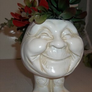 May include: White ceramic planter shaped like a smiling face with a red and green succulent plant arrangement on top. The planter has a whimsical, cartoon-like appearance with exaggerated features.