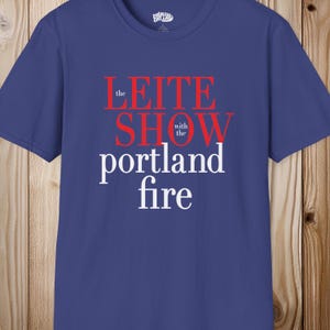 May include: A blue short-sleeved t-shirt featuring the red and white text "the LEITE SHOW with the portland fire" printed on the front. The shirt has a crew neckline and a small brand logo printed on the inside of the collar area.