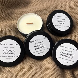 May include: Four round black metal tins containing soy wax candles with wooden wicks. Labels read Spell of Luna, LLC, soy wax candle, 2 oz / 56g, and scents Pumpkin Caramel, Haunted Orchard, Orange Spice, and Cranberry Prosecco, handpoured in Ocala, FL.