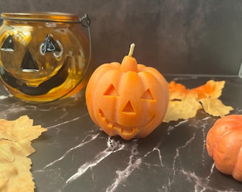 Peut inclure: Une bougie orange en forme de citrouille avec un visage de lanterne sculpté et une mèche unique sur le dessus. La bougie est placée près d'une lanterne en verre en forme de citrouille et de feuilles d'automne artificielles.