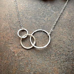 Può includere: Una collana d'argento con tre cerchi interconnessi. La collana è su uno sfondo marrone.