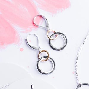 May include: A silver chain necklace with three interlocking rings in silver, rose gold, and dark gray, accompanied by a pair of matching lever-back earrings. A circular card features the text: Storied. Your life is a work in progress.