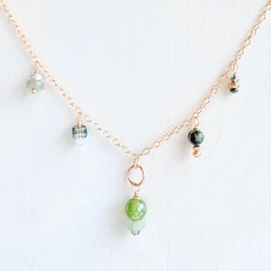 May include: Delicate gold-tone necklace featuring a series of dangling green and blue-green gemstones. The necklace has a simple chain design with a small ring and several gemstone pendants in various shapes and sizes. The gemstones are in shades of green and blue.