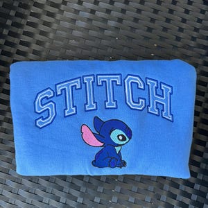 Puede incluir: Sudadera azul con la palabra "STITCH" bordada en blanco y azul. Un personaje azul Stitch con orejas rosas est&aacute; bordado debajo de la palabra.