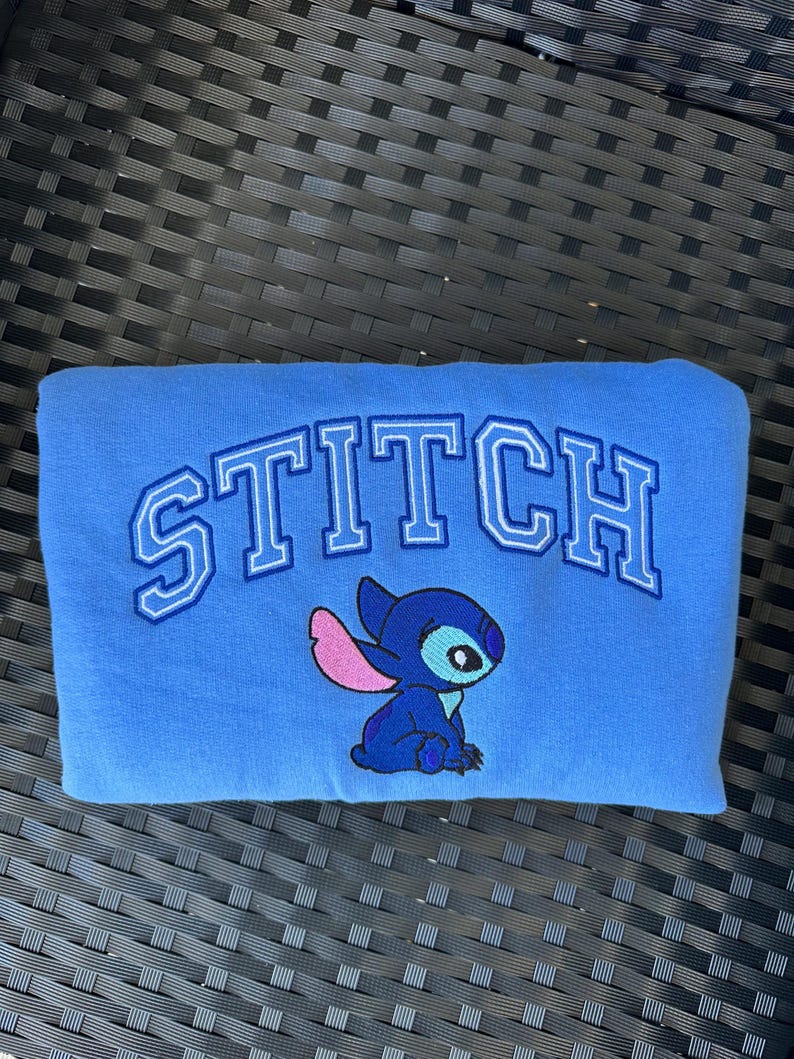 Puede incluir: Sudadera azul con la palabra "STITCH" bordada en blanco y azul. Un personaje azul Stitch con orejas rosas est&aacute; bordado debajo de la palabra.