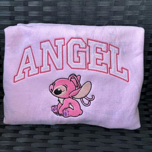 Puede incluir: Sudadera rosa con la palabra "ANGEL" bordada en letras blancas y rojas. Un personaje de dibujos animados rosa con ojos grandes est&aacute; bordado debajo de la palabra.