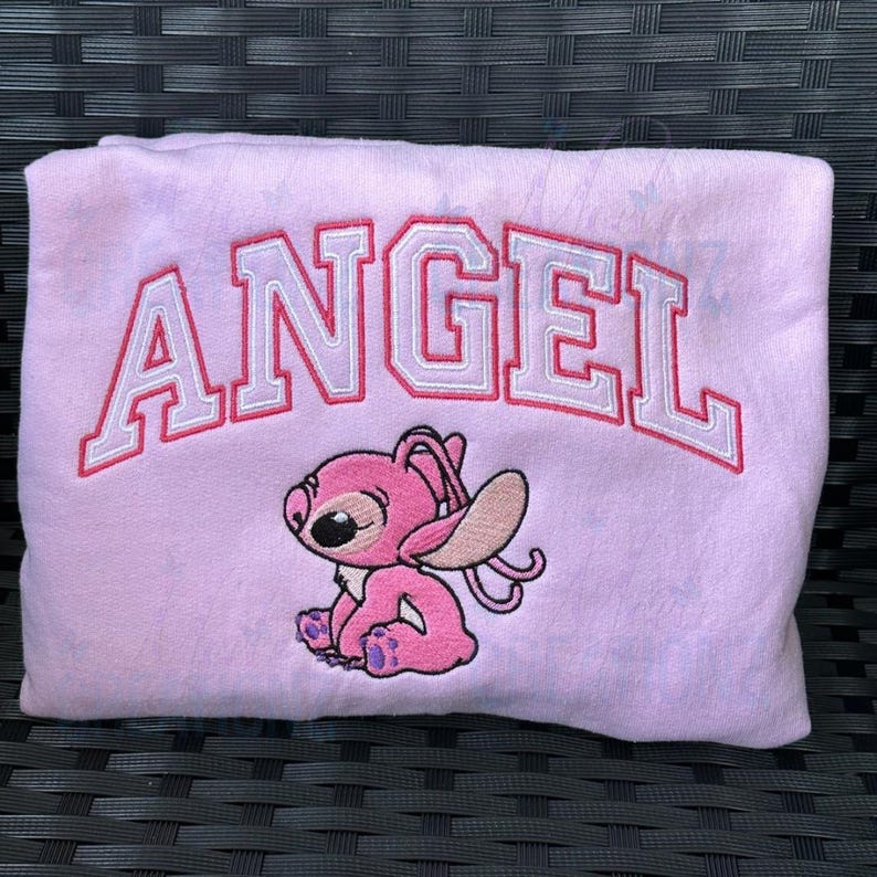 Puede incluir: Sudadera rosa con la palabra "ANGEL" bordada en letras blancas y rojas. Un personaje de dibujos animados rosa con ojos grandes est&aacute; bordado debajo de la palabra.