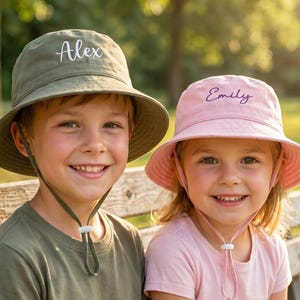 Könnte beinhalten: Zwei Kinder tragen personalisierte Sonnenhüte. Einer ist olivgrün mit dem Namen "Alex" in Weiß gestickt, der andere ist rosa mit "Emily" in Lila. Beide Hüte haben Kinnriemen und sind für den Sonnenschutz konzipiert.