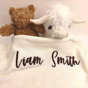 May include: A brown teddy bear and a white stuffed Highland cow rest on a cream-colored knitted blanket. The blanket features the embroidered name "Liam Smith" in a dark brown script font. The toys are set against a white background.