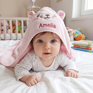May include: A soft, light pink hooded towel featuring embroidered bear ears and the name Amelia stitched in red thread on the front. The hood is designed with a friendly animal face, including eyes, a nose, and a mouth, providing a cozy covering.
