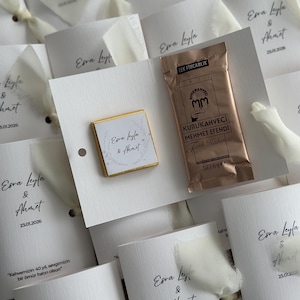 May include: A white card favor featuring a square chocolate with a floral wreath design and the names Esra Leyla & Ahmet, alongside a 6g packet of Kurukahveci Mehmet Efendi Turkish coffee, tied with a white ribbon and printed with the date 23.01.2026.